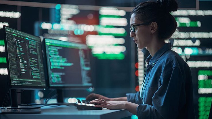 Eine junge Frau mit Brille sitzt in einem Computerraum vor zwei Computerbildschirmen und tippt