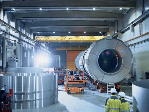 Two people wearing hard hats walk through a production hall for large components
