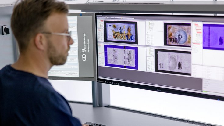 A person is working at a desk with two large computer monitors displaying images of currency notes
