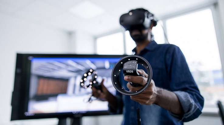 A professional using a virtual reality headset and controllers, standing in an office environment with a monitor displaying a virtual simulation in the background.