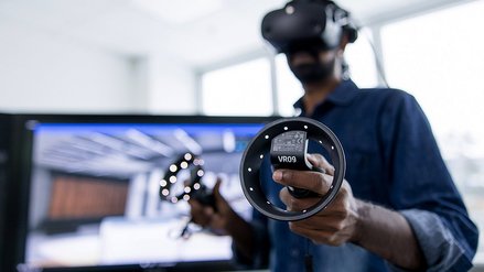 A professional using a virtual reality headset and controllers, standing in an office environment with a monitor displaying a virtual simulation in the background.