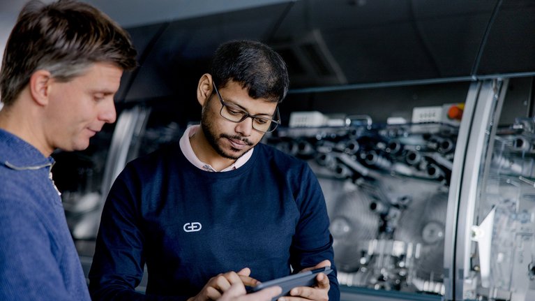 Two professionals in a workshop discussing over a digital tablet, with industrial machinery in the background.