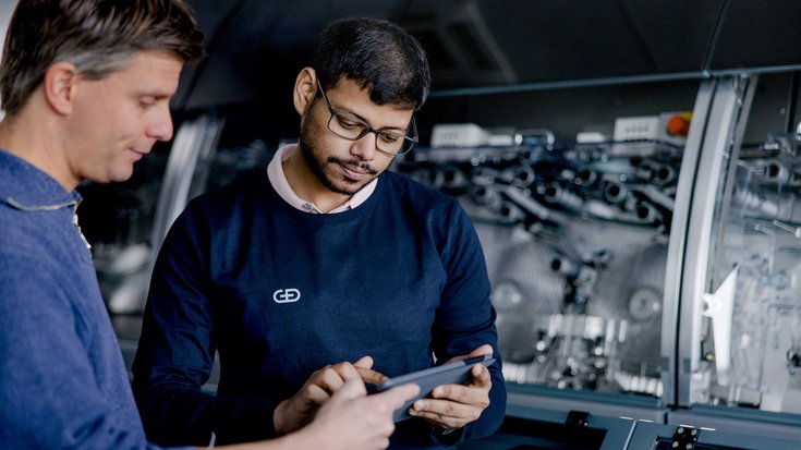 Two professionals in a workshop discussing over a digital tablet, with industrial machinery in the background.