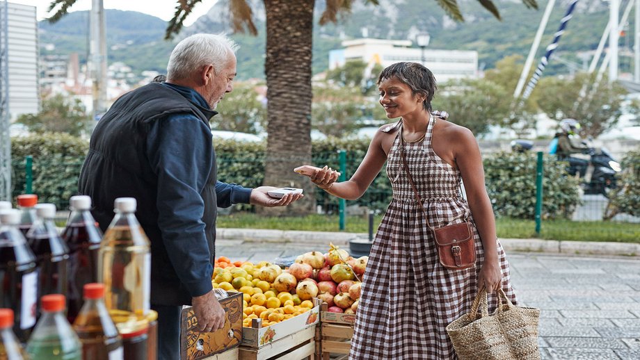 A woman purchasing fruits and vegetables at an outdoor market stall, handing cash to the vendor
