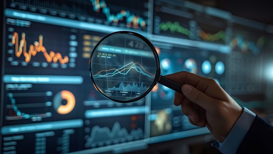 Hand holding a magnifying glass focusing on data charts displayed on multiple screens