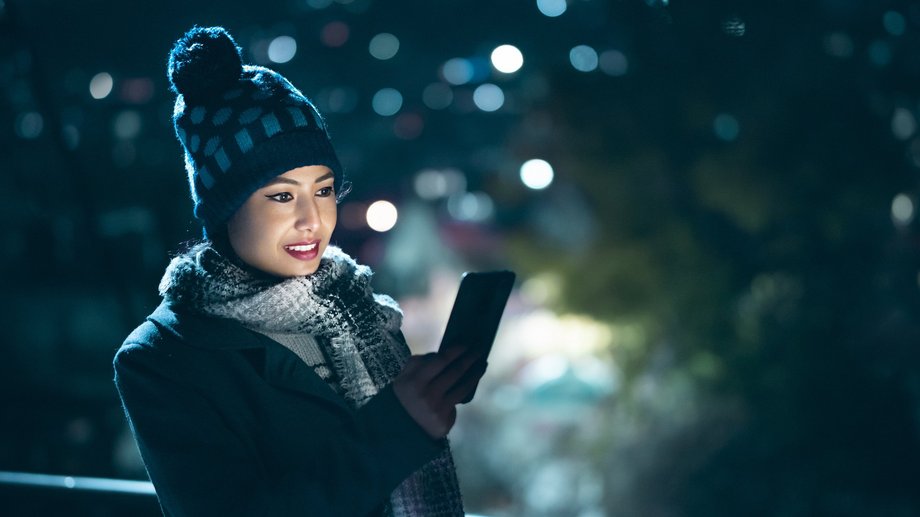 Person in winter attire using a smartphone at night, with blurred city lights in the background