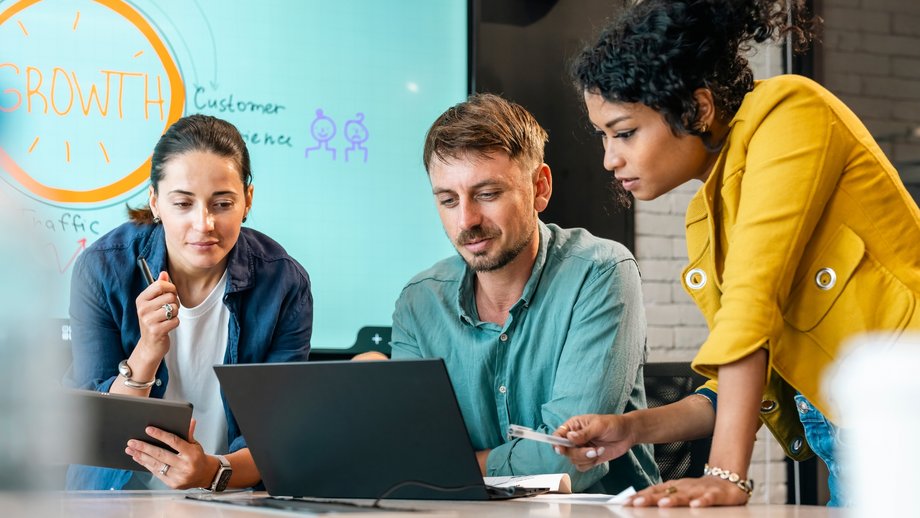 Three people sitting at a board room table looking at a laptop computer in a modern office.