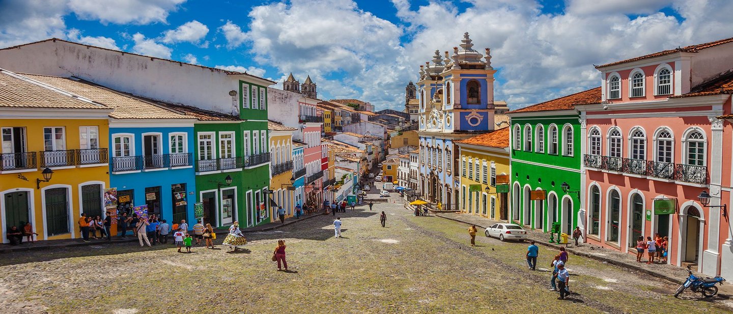 A vibrant street scene in Pelourinho, Salvador, Brazil, with colorful colonial buildings and people walking under a bright blue sky.
