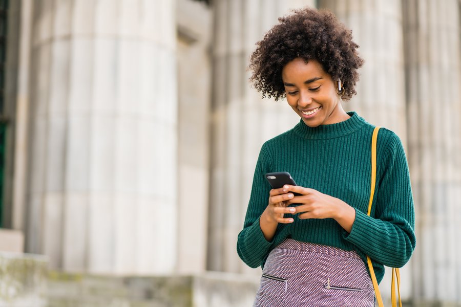  Person in a green sweater standing outside in front of a large pillar and using a smartphone.