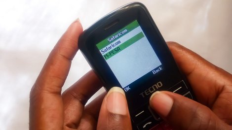 Close-up of a mobile phone showing M-PESA in the menu, held by a person