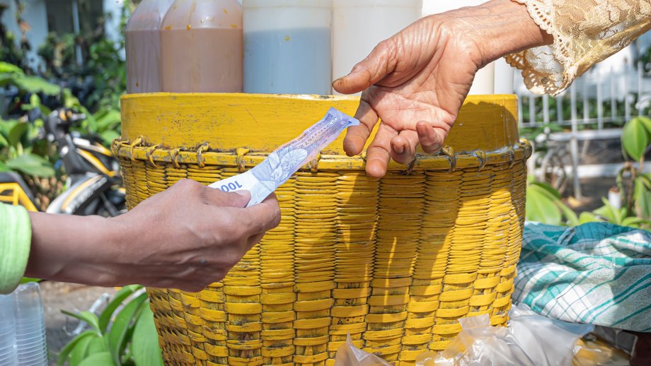 Two hands exchanging an Indonesian Rupiah banknote to pay in front of a woven basket.