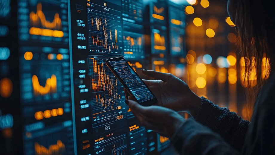 Person using a smartphone to analyze financial data on large screens with graphs and charts