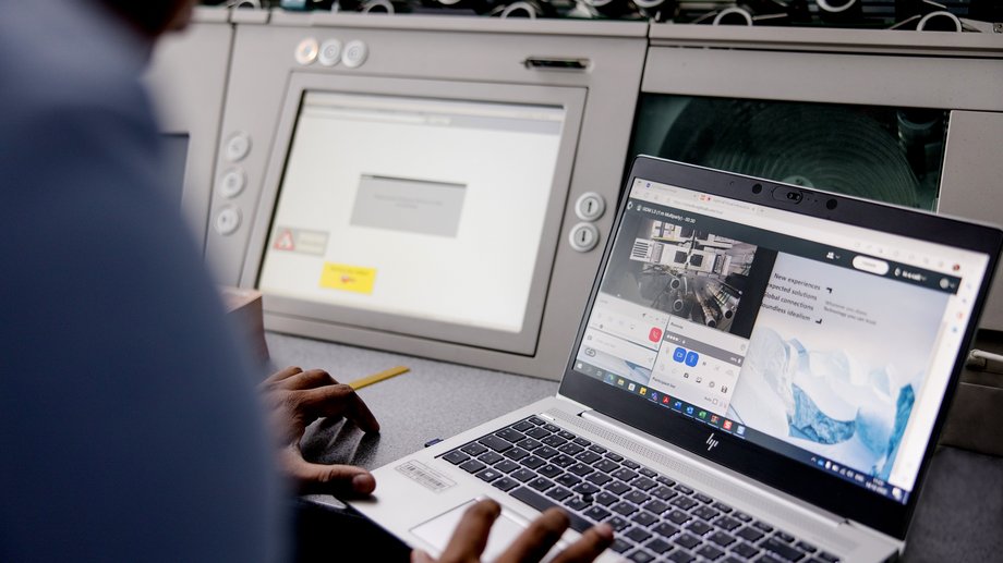 A person using a laptop and control panel, with the laptop displaying a video call and web content.