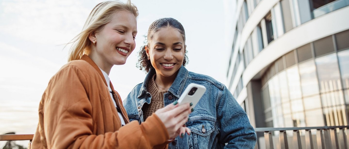 Two young women looking at a white smartphone with futuristic building in the background.