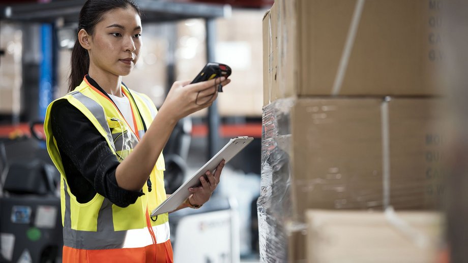 Woman in safety vest scans packages in a warehouse with a handheld scanner and holds a clipboard.