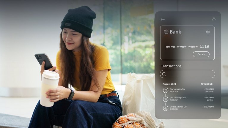 A person seated on a bench holding a coffee cup and a smartphone, viewing a digital banking interface displaying recent transactions.