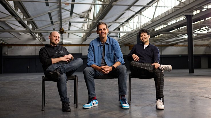 Three men sitting on chairs in a spacious industrial setting, dressed in casual attire.