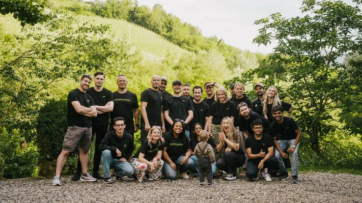 A large group of people, including children, wearing casual clothes and standing in front of a vineyard.
