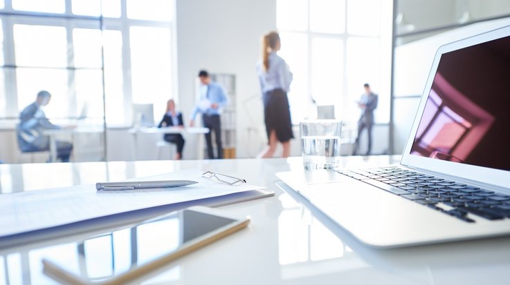On a desk are a clipboard, a pen, a mobile phone, a laptop and a glass of water. An office can be seen in the background.