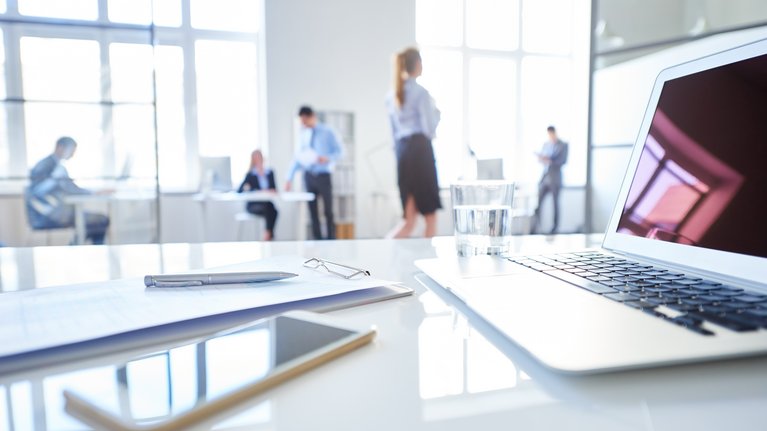 On a desk are a clipboard, a pen, a mobile phone, a laptop and a glass of water. An office can be seen in the background.