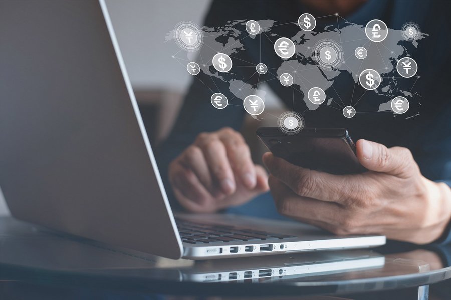 A man in front of a laptop holds a smartphone in his hand, over which floats a digital simulation of a world map with various currency symbols