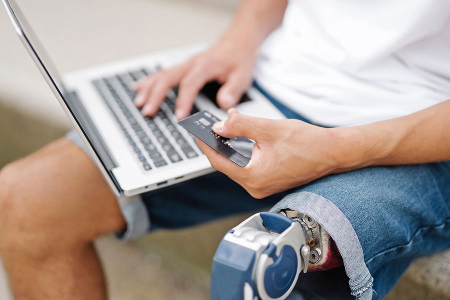 Young man with disability, artificial leg/prosthesis, pays cashlessly with credit card and laptop