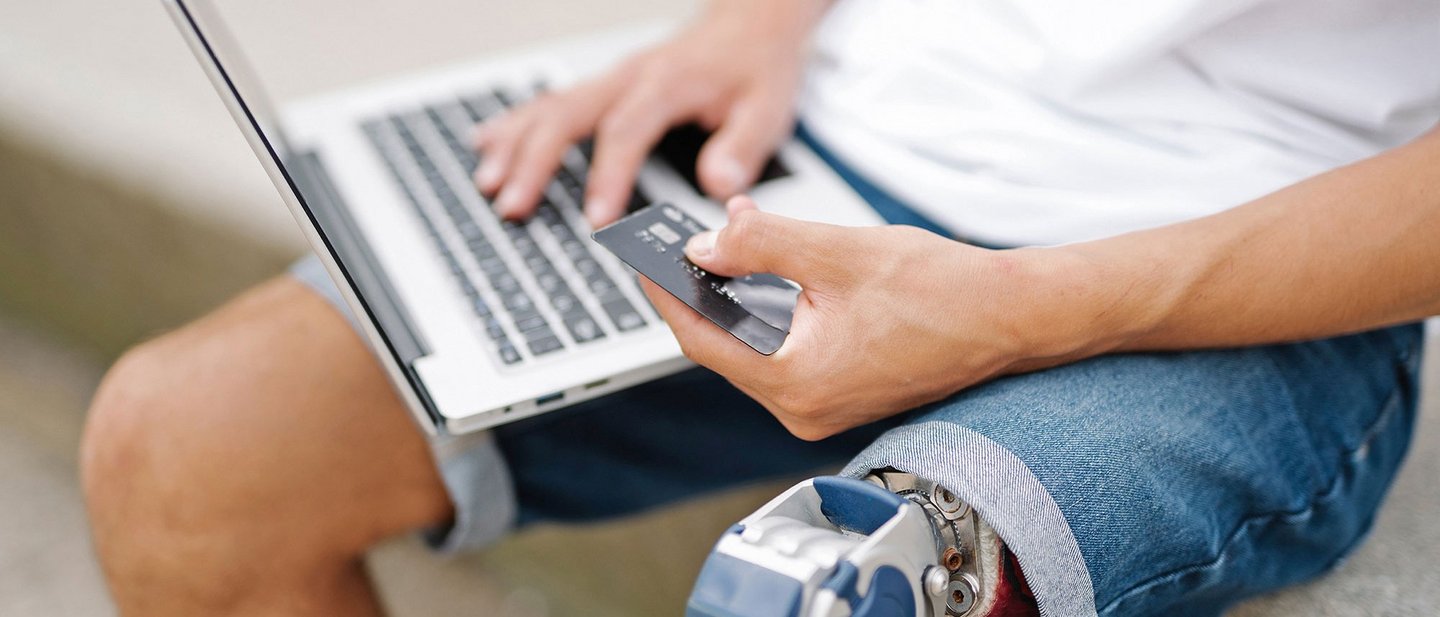 Young man with disability, artificial leg/prosthesis, pays cashlessly with credit card and laptop