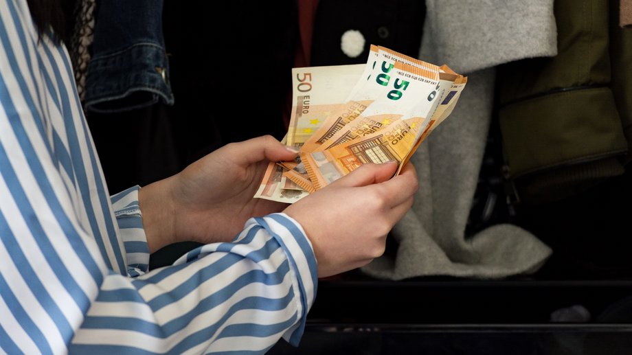 Close-up of hands holding euro banknotes, including €50 and €20, in front of clothing racks.