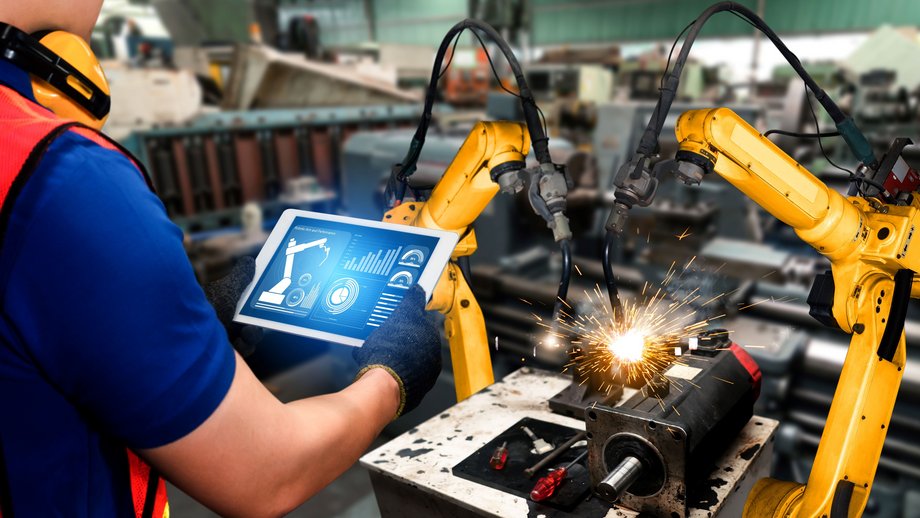 Hand holding a tablet to control a robotic arm in a factory with robotic arms in the background