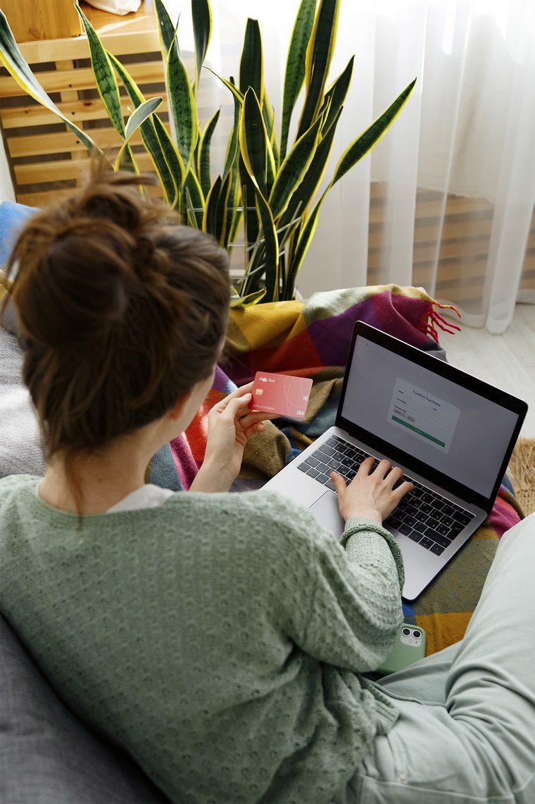 A woman sits on her sofa in front of her laptop and holds a credit card in her hand