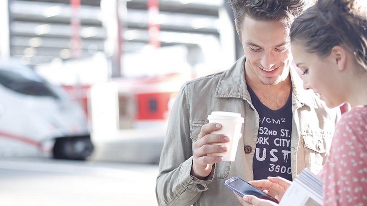 Two young people standing in front of a train station operating a smartphone