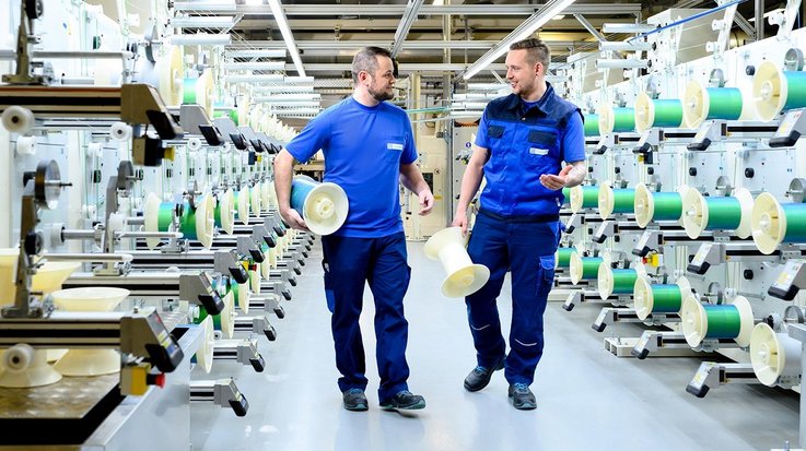 Two men carry coils in a production hall and talk to each other