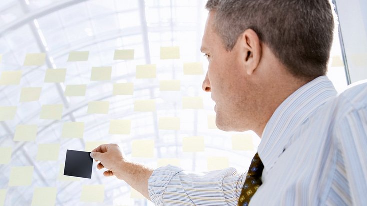 A businessman holds up a black post-it in front of a wall with many yellow post-its
