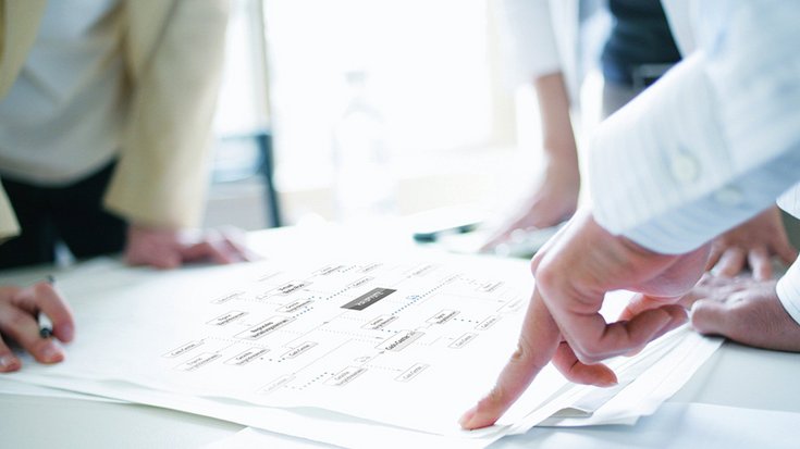 Three people are standing at a table, bent over papers they are discussing