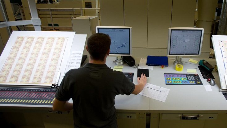 A man stands in front of a sheet of banknotes, next to it are two monitors on a desk