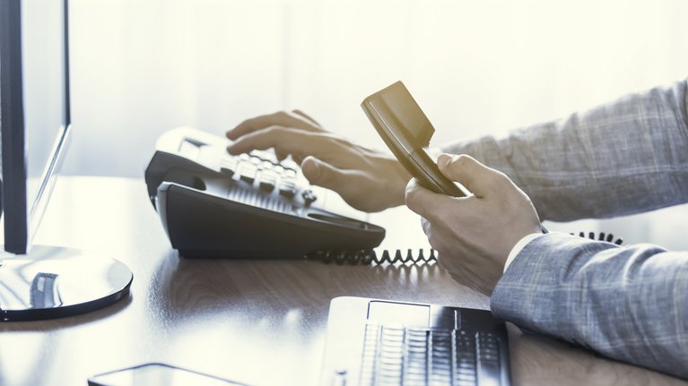 Man sits at a desk, has a telephone receiver in his hand and types in a number