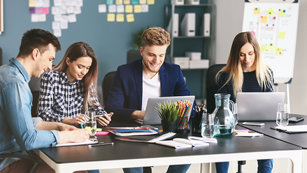Four good-humored people sitting at a desk in an office, working on laptops and paper pads