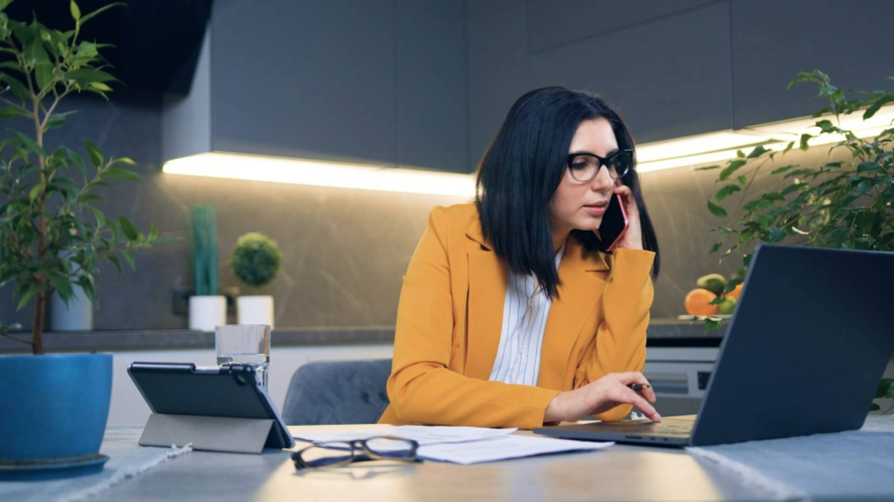A woman sits in her kitchen working remotely with a laptop, tablet and smartphone